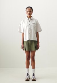 Knappe, kortermede beige skjorte med lommer foran, kombinert med et grønt plisserte skjørt. Hvite sokker og beige sneakers fullfører antrekket.