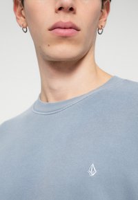 Light blue cotton sweatshirt with a crew neckline, featuring a small white embroidered logo on the left chest and ribbed cuffs.