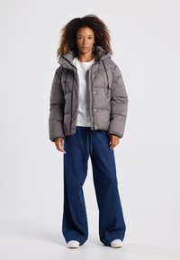Chaqueta acolchada en patrón de cuadros marrón, con un cuello alto y botones de presión. Combinada con pantalones vaqueros sueltos de mezclilla azul oscuro y zapatos blancos.