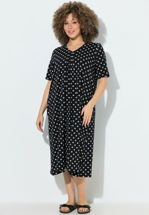 Kobieta z kręconymi włosami, ubrana w czarną sukienkę midi z białymi wzorami gwiazdek i czarne sandały wsuwane, stoi przy jasnej ścianie.