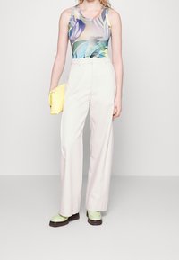 Kobieta w kolorowym topie bez rękawów, białych spodniach o szerokich nogawkach z wysokim stanem, bladozielonych mokasynach i trzymająca żółtą kopertówkę.