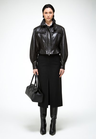 Žena v krátkej čiernej koženej bunde, čiernej midi sukni s predným rozparkom, čiernych čižmách po kolená a s čiernou kabelkou v ruke.