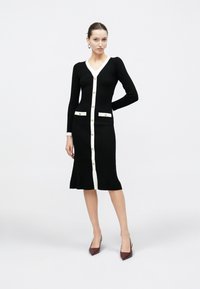 Γυναίκα στέκεται φορώντας μαύρο ριμπ φόρεμα με κουμπιά, με λευκή επένδυση και χρυσά κουμπιά, συνδυασμένο με μπορντό μυτερές γόβες, σε λευκό φόντο.