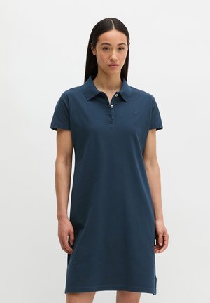 Kobieta w krótkim rękawie granatowej sukience polo stoi na tle jednolitego jasnego tła, zwrócona przodem.