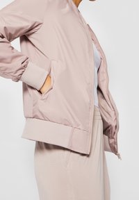 Personne portant une veste bomber rose clair avec des poches sur une chemise blanche et un pantalon beige, debout avec une main dans la poche.