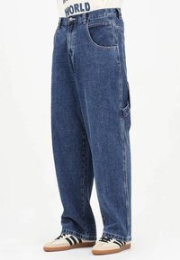 Jeans in denim azzurro a gamba larga con vita alta, tessuto resistente, tasche frontali e laterali, e dettagli di cuciture sottili. Abbinati a sneakers beige.