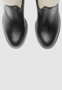 Un par de botas de cuero negro vistas desde arriba sobre un fondo claro y liso, mostrando puntas redondeadas y detalles de costura.