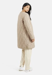 Cappotto lungo beige trapuntato con scollatura rotonda e spacchi laterali, indossato sopra pantaloni bianco sporco e sneakers bianche. Presenta una superficie testurizzata.