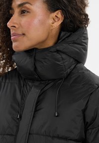 Svart puffjacka med hög krage, med dragsko, tryckknappar och en glansig yta. Närbild av en kvinnas profil.