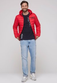 Rote Pufferjacke mit Kapuze und Reißverschluss, mit einem Logo auf der Brust. Über einem dunkelgrauen T-Shirt und hellblauen Jeans getragen. Weiße Sneakers.