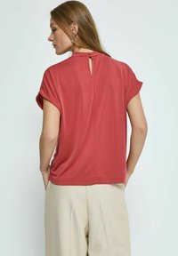 Raudona trumpomis rankovėmis palaidinė su nugaros rakteliu, pagaminta iš minkštos medžiagos. Dėvima su šviesiai bežinėmis aukšto liemens kelnėmis.