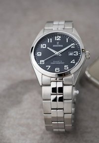 Montre-bracelet en acier inoxydable argenté avec un cadran analogique noir, de grands chiffres blancs, un affichage de la date et un bracelet en maillons métalliques.