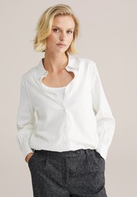 Camicia bianca in cotone con scollatura a V e dettagli arricciati sulle maniche, abbinata a pantaloni grigio scuro con texture.