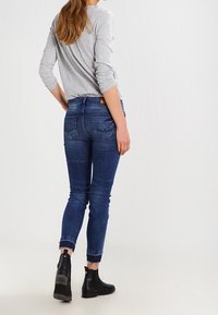 Džínové kalhoty v tmavě modré barvě se slim fit a roztrhaným lemem, spárované se světle šedou košilí s dlouhým rukávem a černými kotníkovými botami.