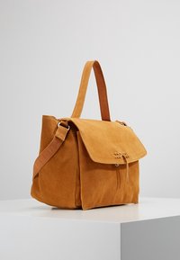 Sac à main en daim marron clair avec une fermeture à rabat, une bandoulière en cuir réglable et un détail de couture décorative à l'avant. Forme carrée avec une texture douce.