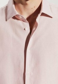 Camisa de botones rosa claro con cuello de solapa, que presenta una textura suave y un ligero brillo. Dos botones visibles en el cuello.