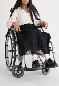 Une personne en fauteuil roulant porte un chemisier crème clair et une jupe plissée noire. Des chaussures noires et des chaussettes blanches complètent la tenue.
