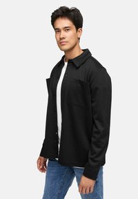 Svart knappet jakke med to frontlommer, laget av et glatt stoff. Båret over en hvit skjorte, paret med blå jeans.