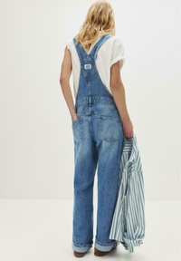 Persoon met blond haar in blauwe denim overalls en een wit t-shirt, die een groen en wit gestreept shirt vasthoudt tegen een neutrale achtergrond.