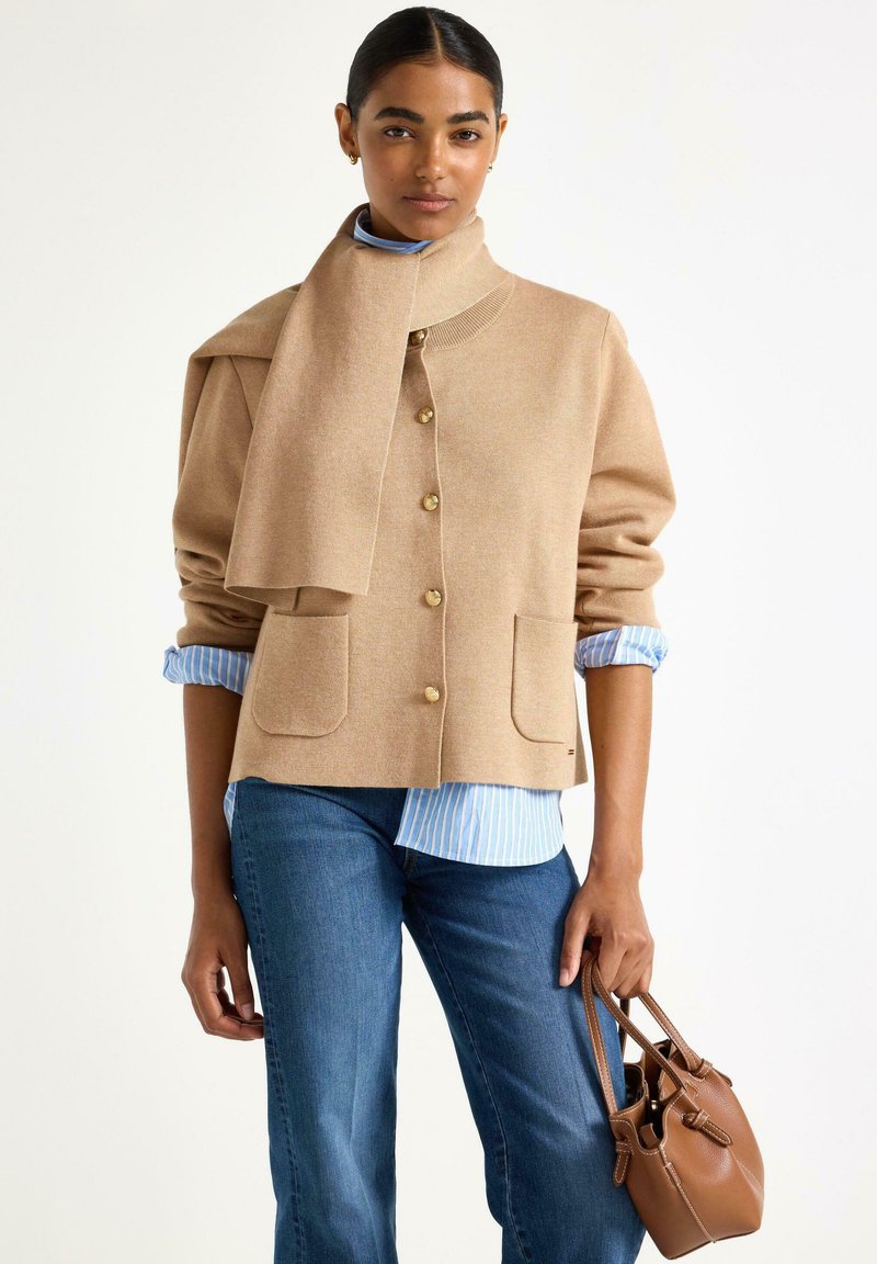 Cardigan in lana beige con colletto alto, bottoni dorati e due tasche frontali. Indossato sopra una camicia a righe blu e abbinato a jeans in denim.