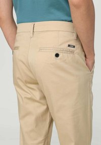 Pantaloni beige con una texture liscia, dotati di due tasche posteriori e un piccolo etichetta di branding vicino alla tasca destra.