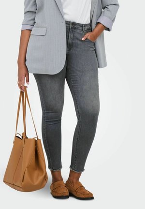 Žena oblečená v sivých úzkych džínsoch, svetlosivom saku, bielom tričku, hnedých mokasínach, ktorá drží veľkú koženú tašku hnedej farby.