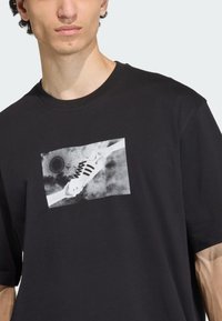 Čierne bavlnené tričko s šedou grafikou topánky na bielom pozadí v strede. Krátke rukávy a voľný strih.