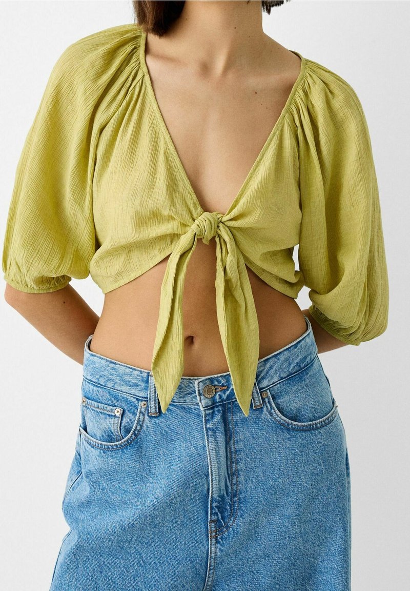 Top corto texturizzato verde chiaro con maniche a sbuffo e nodo frontale. Abbinato a pantaloni corti in denim azzurro a vita alta.