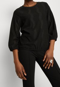 Blusa nera con maniche a tre quarti, tessuto testurizzato con motivi a onde intagliati e scollatura tonda.