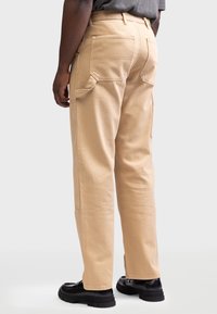 Beige cargo bukser med en indsnævret ben design, der har flere lommer og et robust bomulds materiale. Bæres med sorte støvler.