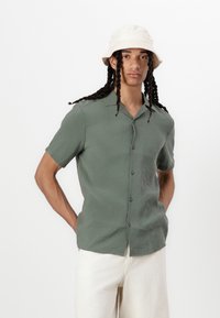 Green short-sleeve button-up shirt in a lightweight fabric, with a relaxed fit and a flat collar, worn with off-white pants and a beige bucket hat.