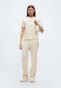 Μπλούζα κρεμ χρώματος με το κείμενο «Calvin Klein Jeans», συνδυασμένη με τζόγκερ στο ίδιο κρεμ χρώμα και μικρή τσάντα, όλα από απαλό ύφασμα. Άσπρα αθλητικά παπούτσια.