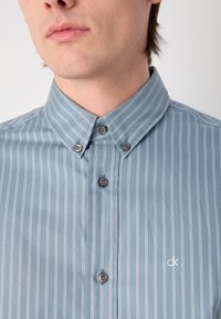 Cămașă bleu deschis, cu mânecă scurtă, cu nasturi, având dungi verticale albe înguste, guler ascuțit și logo brodat pe piept.