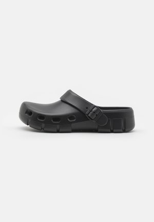 Zoccolo nero slip-on con cinturino regolabile, fori di ventilazione ai lati e suola spessa, mostrato su sfondo bianco.