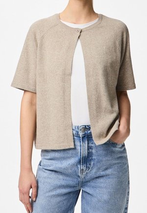 Person iført en kortærmet beige strikket cardigan knappet øverst over en hvid skjorte, kombineret med højtaljede blå jeans med en hånd i lommen.