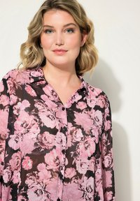 Blumenbluse aus durchsichtigem schwarzem Stoff mit rosa Rosen. Klassischer Kragen und Knopfleiste vorne mit langen Ärmeln. Subtile Textur, elegantes Design.