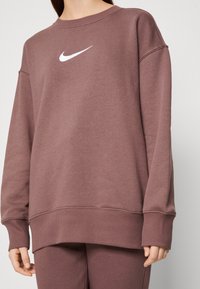 Sudadera púrpura con cuello redondo, puños y dobladillo acanalados, y un logo de Nike blanco centrado en el pecho. Tela suave y texturada.