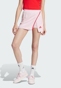 Svetlo roza športni kratek hlačnik z elastiko v pasu, rdečim zadrgo in Adidas logotipom na strani, v kombinaciji z belimi kratkimi nogavicami in roza športnimi čevlji.