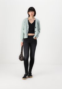 Ljus mintgrön stickad kofta med knappar, svart kort topp, svarta smala jeans och svarta sneakers med vita ränder, bärande en mörk väska.