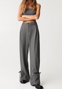 Pantalons larges gris avec une texture de tissu, fermeture à bouton et accents à cordon au niveau de l'ourlet. Associés à un haut sans bretelles assorti.