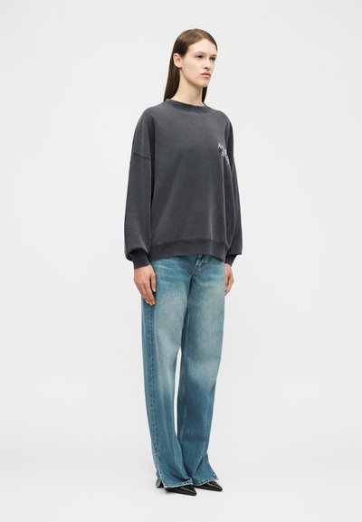 Jauna moteris stovi, vilkėdama laisvus išblukusius mėlynus džinsus, per didelį tamsiai pilką džemperį ir juodus aštrius batelius priešais baltą, paprastą fono sieną.