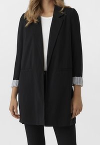 Woman wearing a black blazer with rolled-up pinstriped cuffs over a white shirt and black pants, shown from neck to mid-thigh.