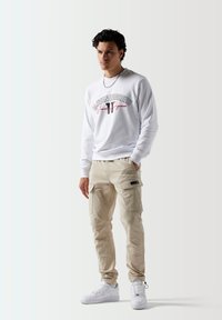 Camisa blanca de manga larga con escritura negra y roja, combinada con pantalones cargo beige y zapatillas blancas. Detalles de bolsillos notables en los pantalones.