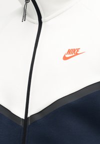 Veste zippée blanche et marine avec une fermeture éclair noire et un logo Nike orange sur la poitrine supérieure.