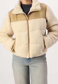 Kremowa bluza polarowa z beżowym nylonowym panelem na ramionach i kołnierzu, z zamkiem błyskawicznym z przodu i bocznymi kieszeniami. Materiał o teksturze pluszu.