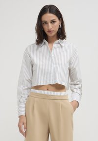 Camicetta accorciata con strisce verticali bianche e dorate, con bottoni davanti e colletto a punta. Abbinata a pantaloni beige a vita alta.