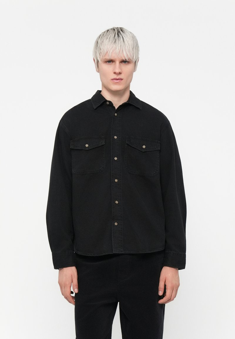 Black shirt featuring a button-up design, two chest pockets, and long sleeves. Made of cotton with a smooth texture.