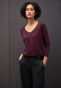 Maglione bordeaux con scollo a V e maniche aderenti, abbinato a pantaloni grigio scuro su misura con tasche laterali e chiusura con bottone.
