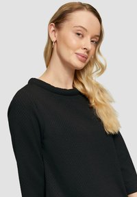 Zwarte textuurtrui met een ronde halslijn en 3/4 mouwen. Het model heeft lang, golvend haar en gouden creolen.