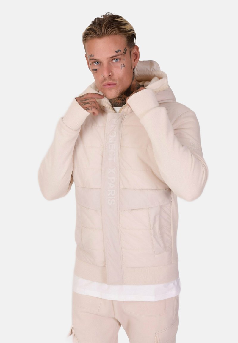 Haut à capuche beige clair avec rembourrage matelassé, manches longues et fermeture éclair sur le devant. Présente le logo "PROJECT X PARIS" sur la poitrine.